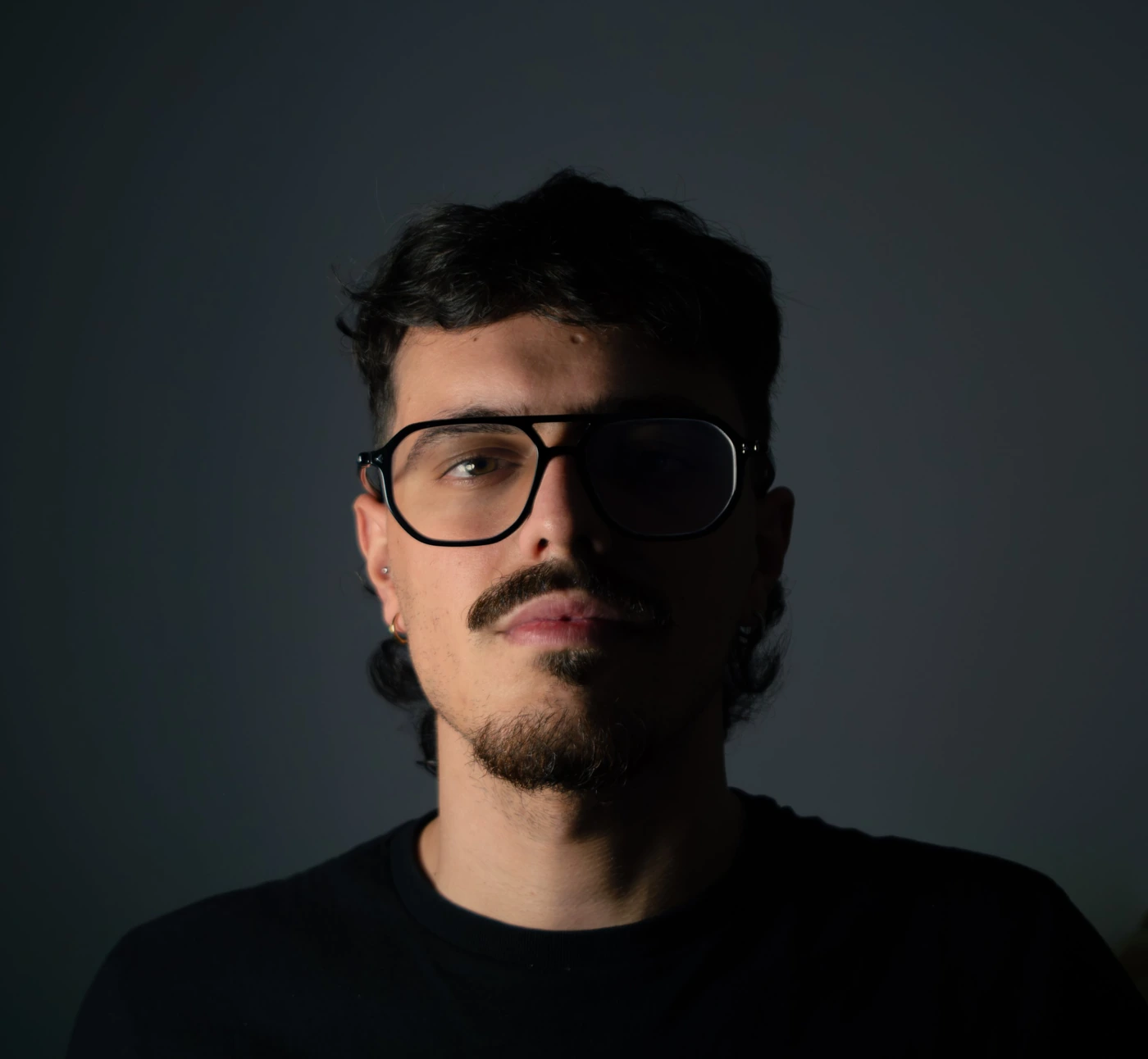 Retrato de Juan Salvador con gafas de pasta negras y camiseta negra, iluminado de lado sobre un fondo gris oscuro