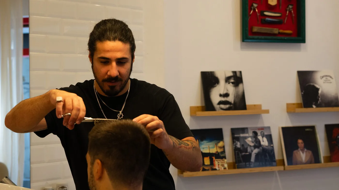 Barbero con camiseta negra cortando el pelo con tijera y peine, junto a un cuadro de herramientas antiguas y discos en la pared